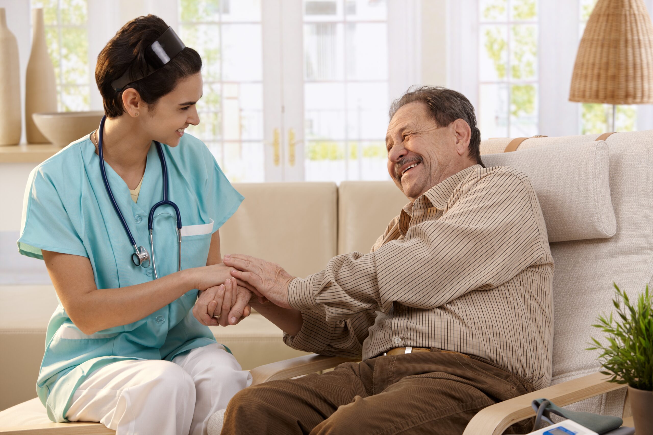 Happy nurse holding hands of elderly patient sitting side by side at home, laughing.