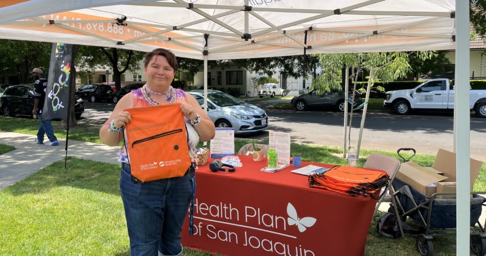 A member from the community smiling and holding up an orange Health Plan of San Joaquin/Mountain Valley Health Plan drawstring bag. They are standing in front of a Health Plan of San Joaquin booth outdoors.