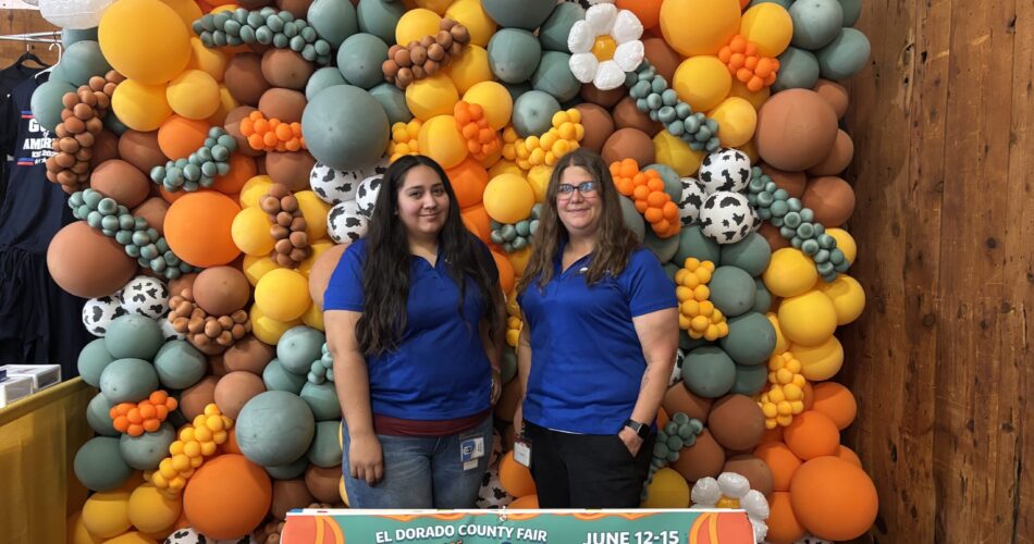 2 employees from Health Plan of San Joaquin posing in front of a balloon backdrop at the 2025 El Dorado County Moovin 'n Groovin Event.