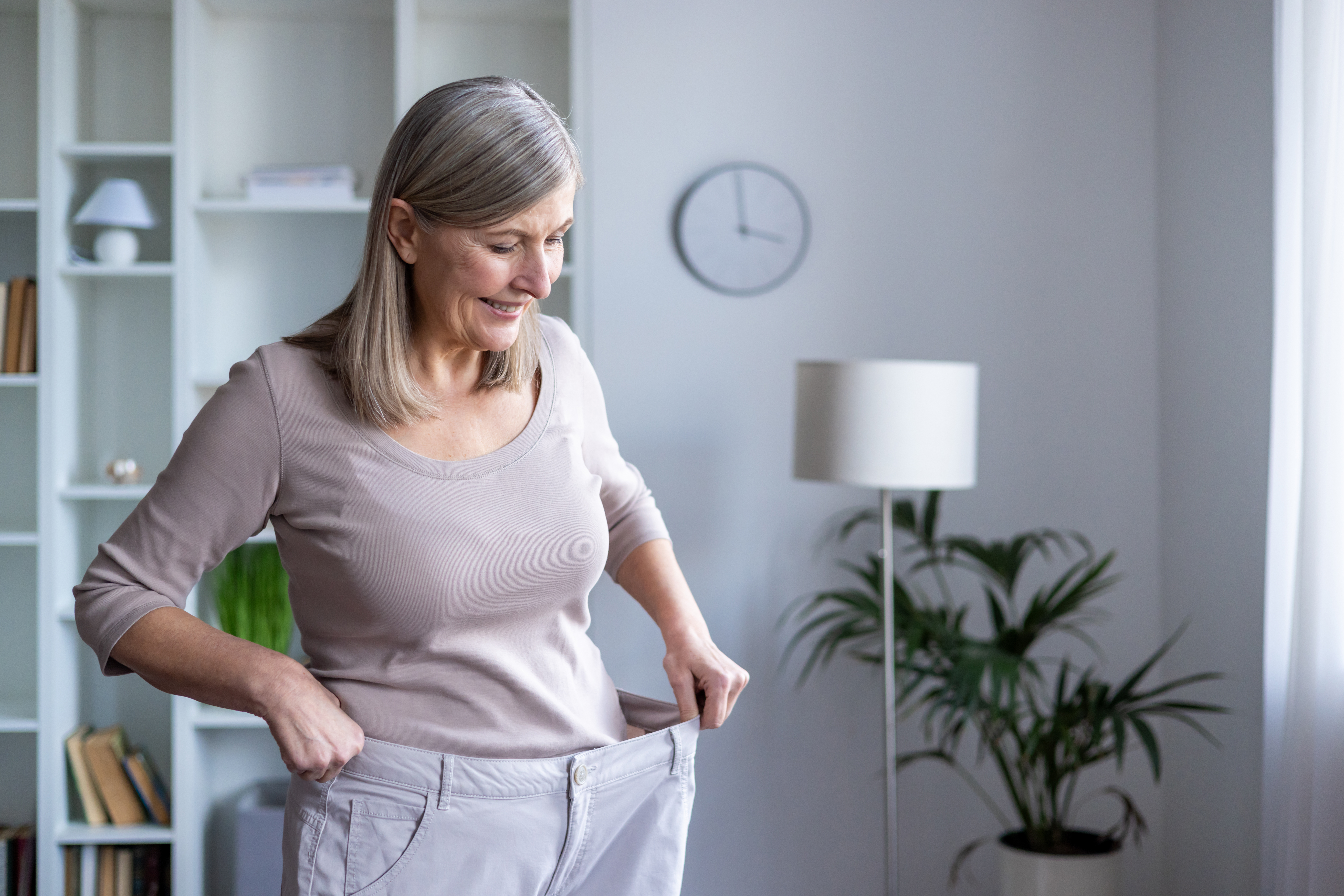 Smiling senior woman feels accomplished as she admires her weight loss progress in her large pants, standing in a cozy room, symbolizing healthy lifestyle and personal achievement. An older woman smiling as she noticed she has lost weight